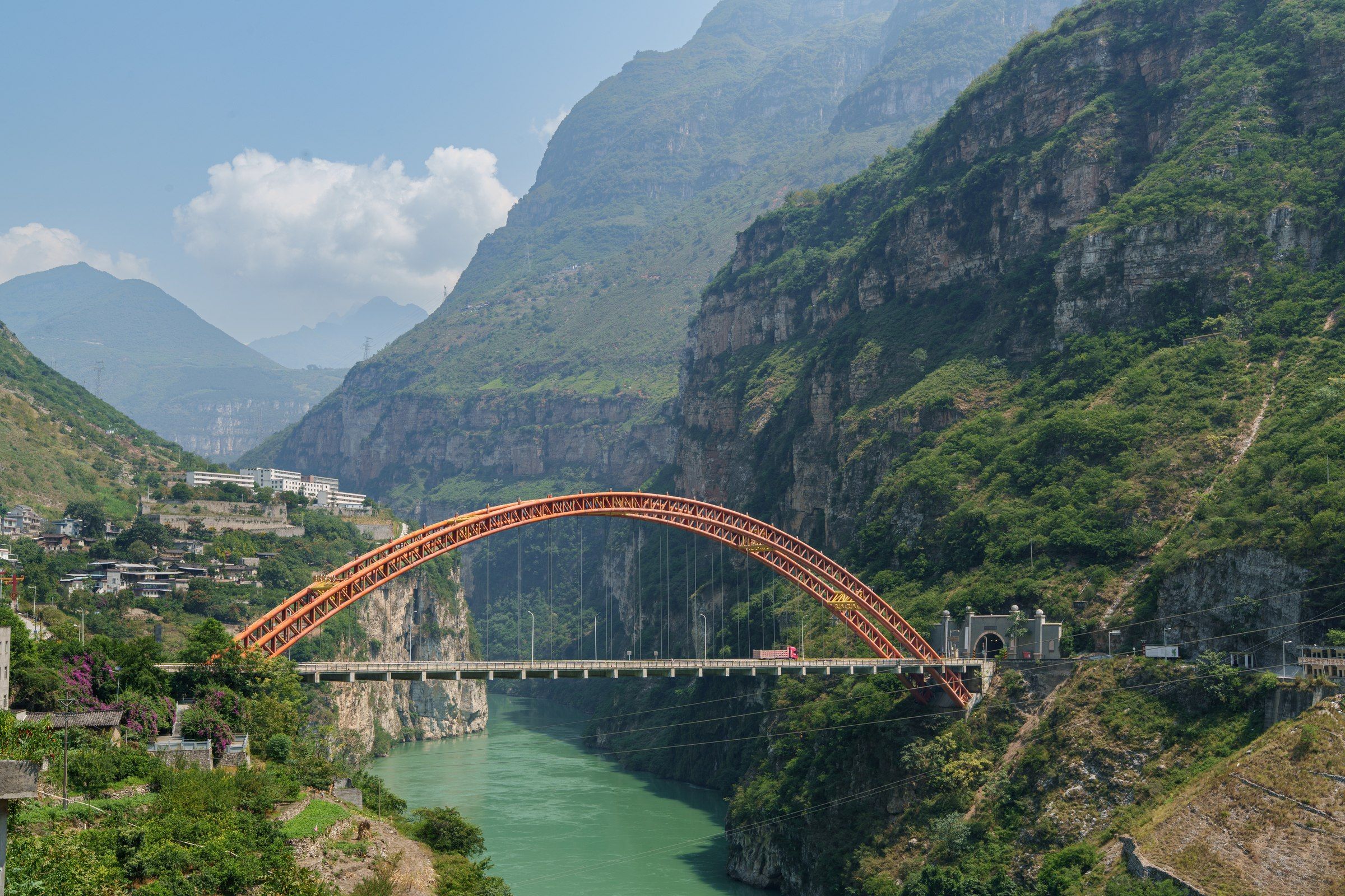 红色拱桥跨过峡谷中的碧绿河水，远处层叠着山峰
