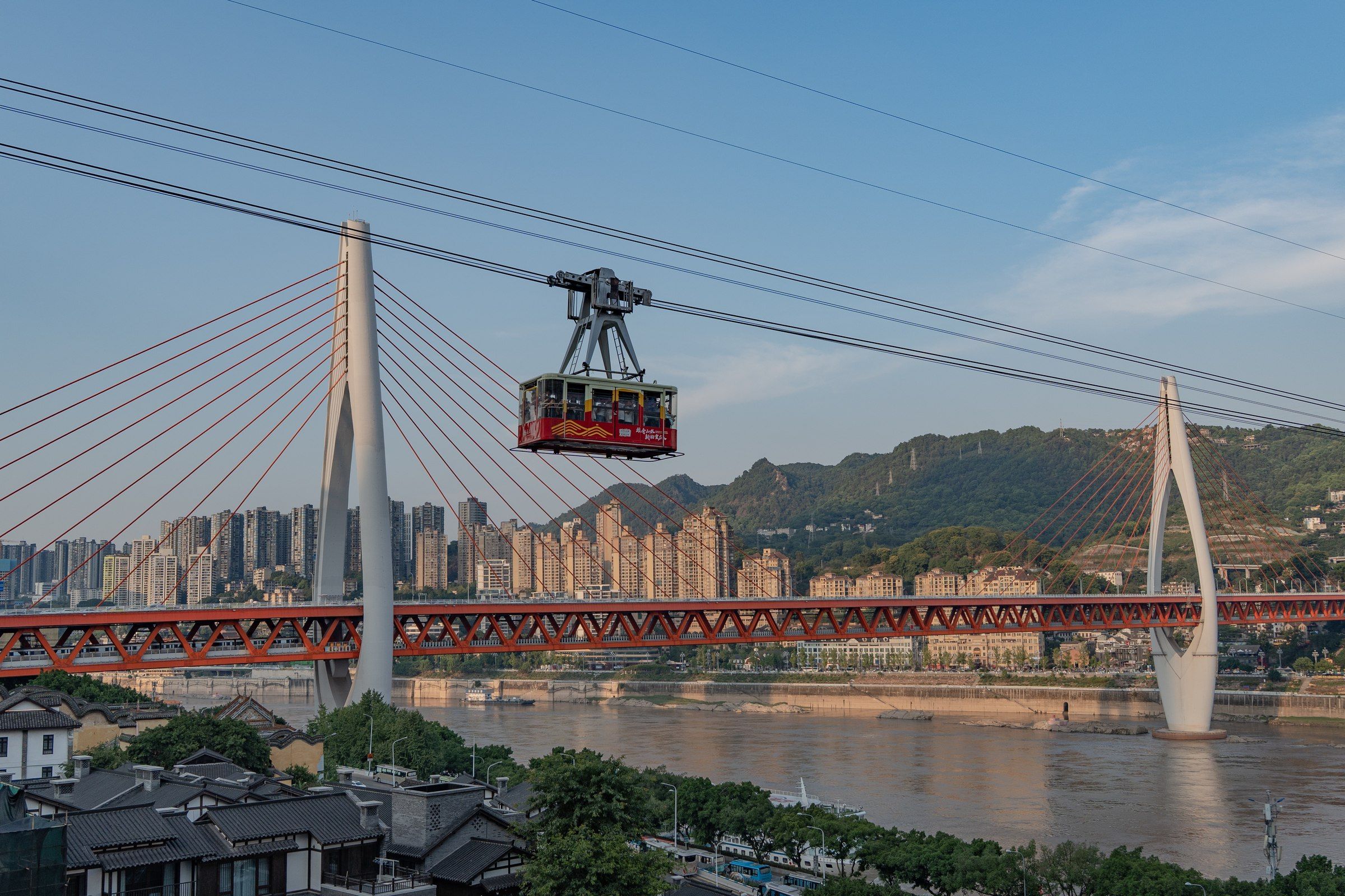 红色索道车厢悬在河上，背后是斜拉桥与山坡